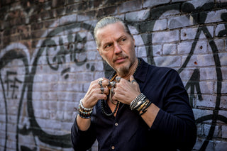 caucasian male model leaning against a graffiti wall with a purple cardigan on while displaying an assortment of men's jewelry accessories