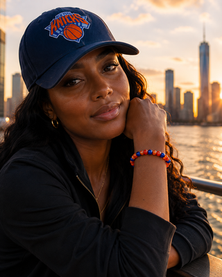Woman wearing a navy blue cap with a logo, sitting by a waterfront with city skyline at sunset.
