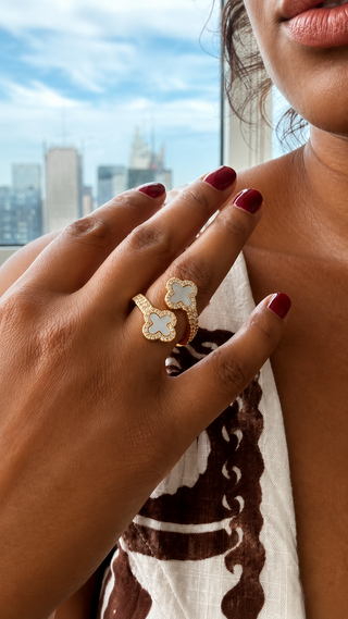 A close-up lifestyle shot of a woman's hand wearing a gold ring with two interlocking clover shapes set with light blue and white stones. The background shows a blurred city skyline and a window view.