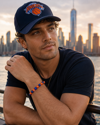 Man wearing a navy blue cap with a logo and a dark t-shirt, sitting by a waterfront with a city skyline in the background.