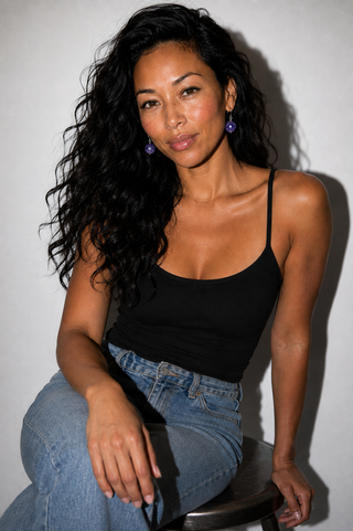 A woman with long, wavy black hair sits on a stool wearing a black spaghetti-strap tank top and light blue jeans against a plain light gray wall.