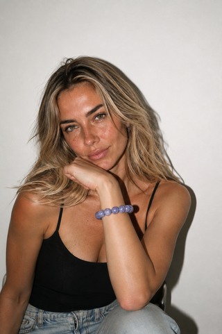 A woman with blonde-highlighted hair wearing a black camisole top and light-wash jeans, resting her chin on her hand to showcase a lavender-purple beaded bracelet on her wrist, set against a plain off-white wall.