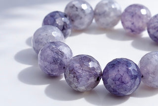 Faceted round gemstone beads in shades of lavender purple and white, arranged in a circle on a white surface with soft shadows.