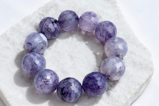 A beaded stretch bracelet made of faceted purple and lavender gemstone beads, displayed on a white stone slab.