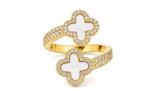 A gold-plated double ring featuring two clover motifs with white inlay and surrounded by small clear pavé stones on the bands, shown on a plain white background.