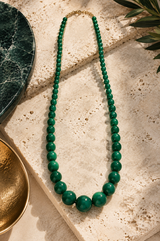 A long strand necklace made of glossy green beads with a small gold clasp, arranged on a textured beige stone surface with decorative green leaves and a gold bowl nearby.