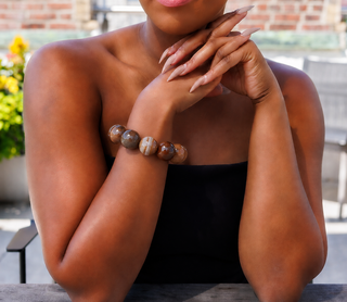 A close-up lifestyle shot of a woman wearing a beaded bracelet made of polished brown and gray stone beads, posed outdoors with her hands crossed.