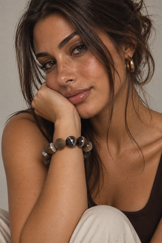 A woman resting her head on her hand, wearing a beaded bracelet made of polished brown and gray stone-like beads, along with gold hoop earrings.