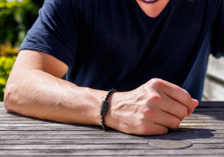 A close-up of a person's forearm resting on a wooden table outdoors, wearing a dark beaded bracelet with a central dark stone.
