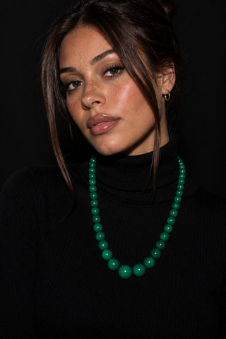 A woman wearing a vibrant green beaded necklace with matching green drop earrings, styled with a black turtleneck against a dark background.